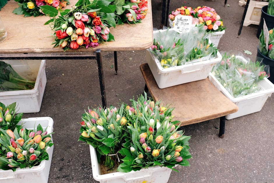 A display of fresh tulip bouquets arranged in white plastic containers and on wooden tables at a flower market. The tulips feature a variety of colors including pink, yellow, white, and purple, with s