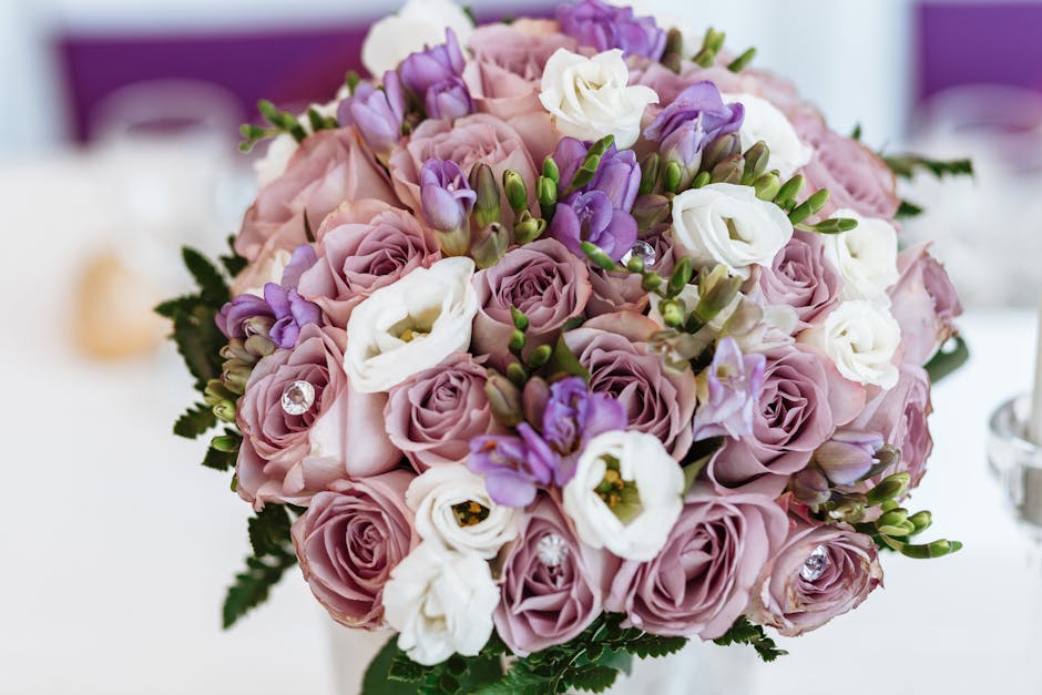 A close-up of a floral bouquet arranged with soft pink roses, white lisianthus, and purple freesias, accented with green foliage. The flowers are fresh, with velvety petals and sturdy stems, carefully