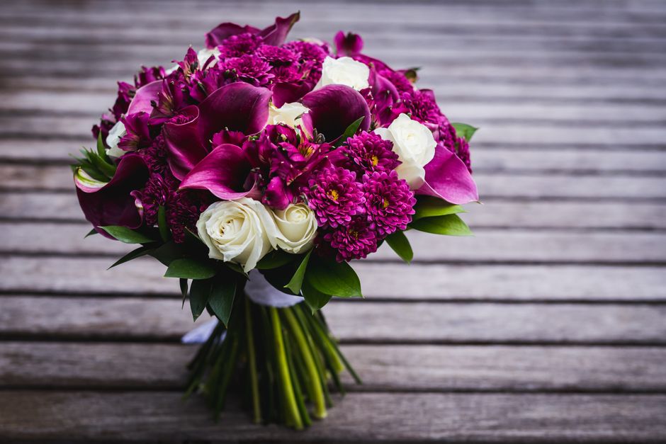 A vibrant floral bouquet featuring deep magenta calla lilies, bright pink chrysanthemums, and purple orchids, complemented by white roses and lush green foliage. The arrangement is hand-tied with visi