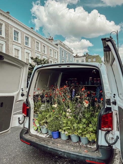 The back of a white delivery van open to reveal an array of potted plants and vibrant flower stems, including red and white blossoms, arranged on shelves and on the floor inside. The flowers appear fr