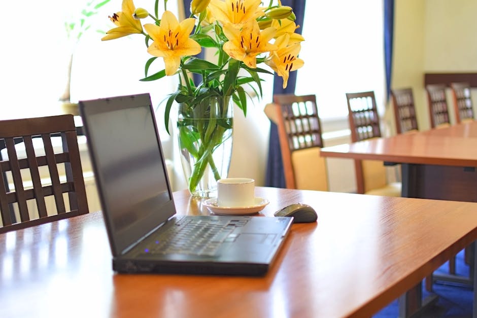 A clear glass vase filled with vibrant yellow lilies with orange streaks and dark stamens, placed on a polished wooden conference table. The bouquet is arranged with lush green leaves and stems, empha