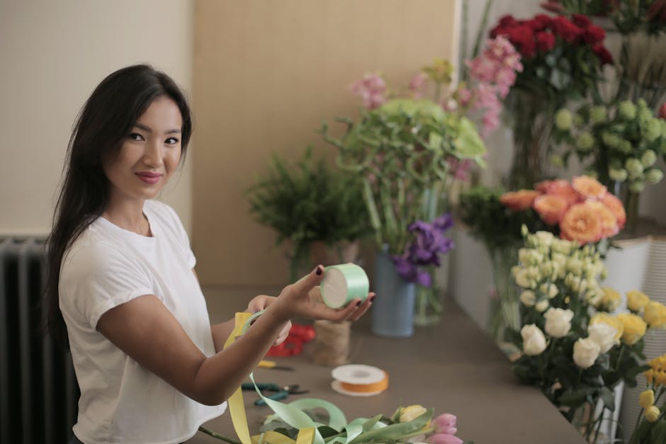 A female florist with long dark hair, dressed in a white t-shirt, is holding a roll of light green floral tape while smiling slightly. She is in a well-lit workspace with a beige wall background, surr